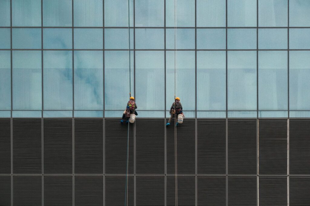 A rope access technician in safety gear cleans the exterior windows of a modern residential high-rise building in Melbou