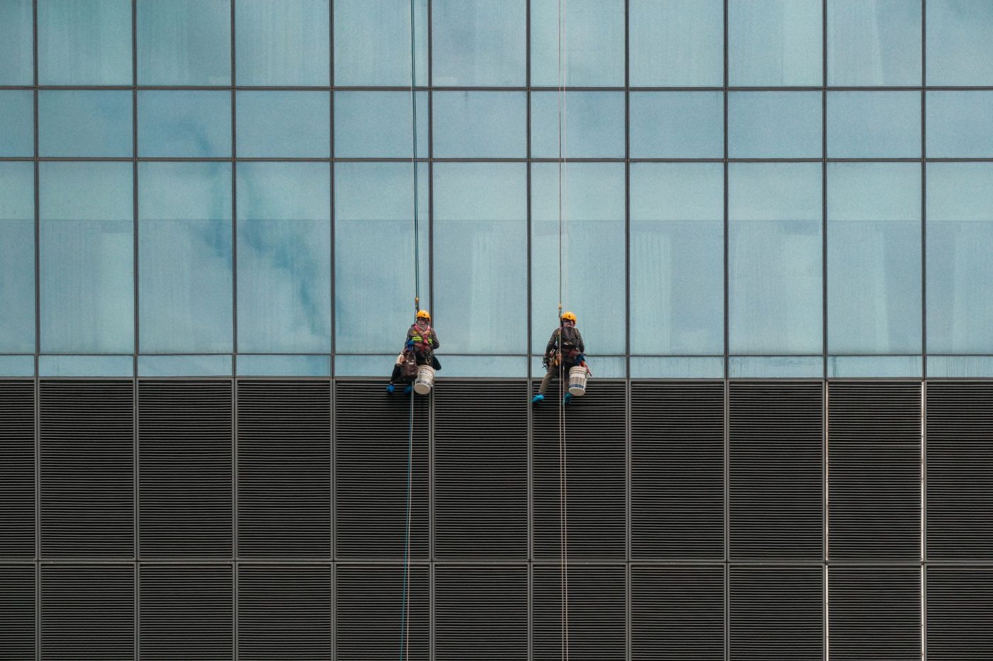 A rope access technician in safety gear cleans the exterior windows of a modern residential high-rise building in Melbou