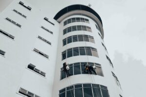A rope access technician in a harness and helmet clears wet leaves from a gutter on a multi-storey residential building,